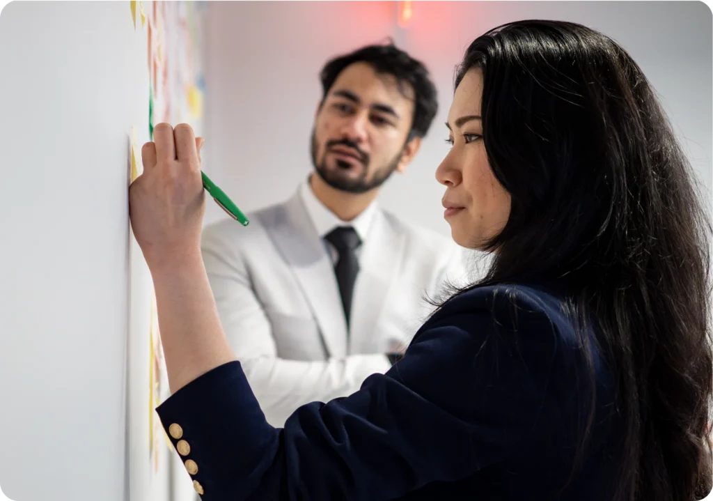 A woman writing on sticky notes on a wall with a green marker while a man in a light suit watches