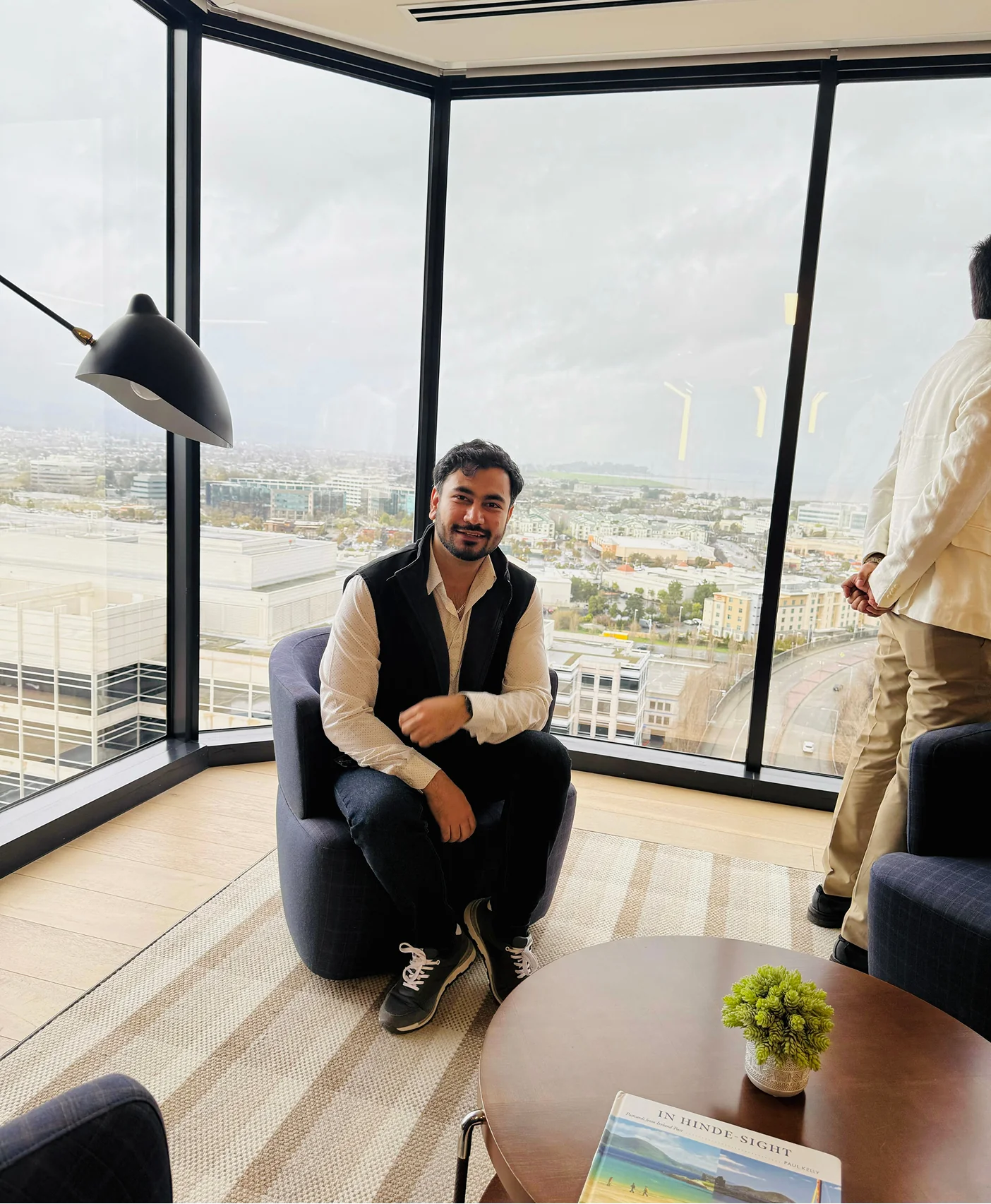 faqs A man sitting in a chair in front of a large window with a city view, a book on a table in front of him