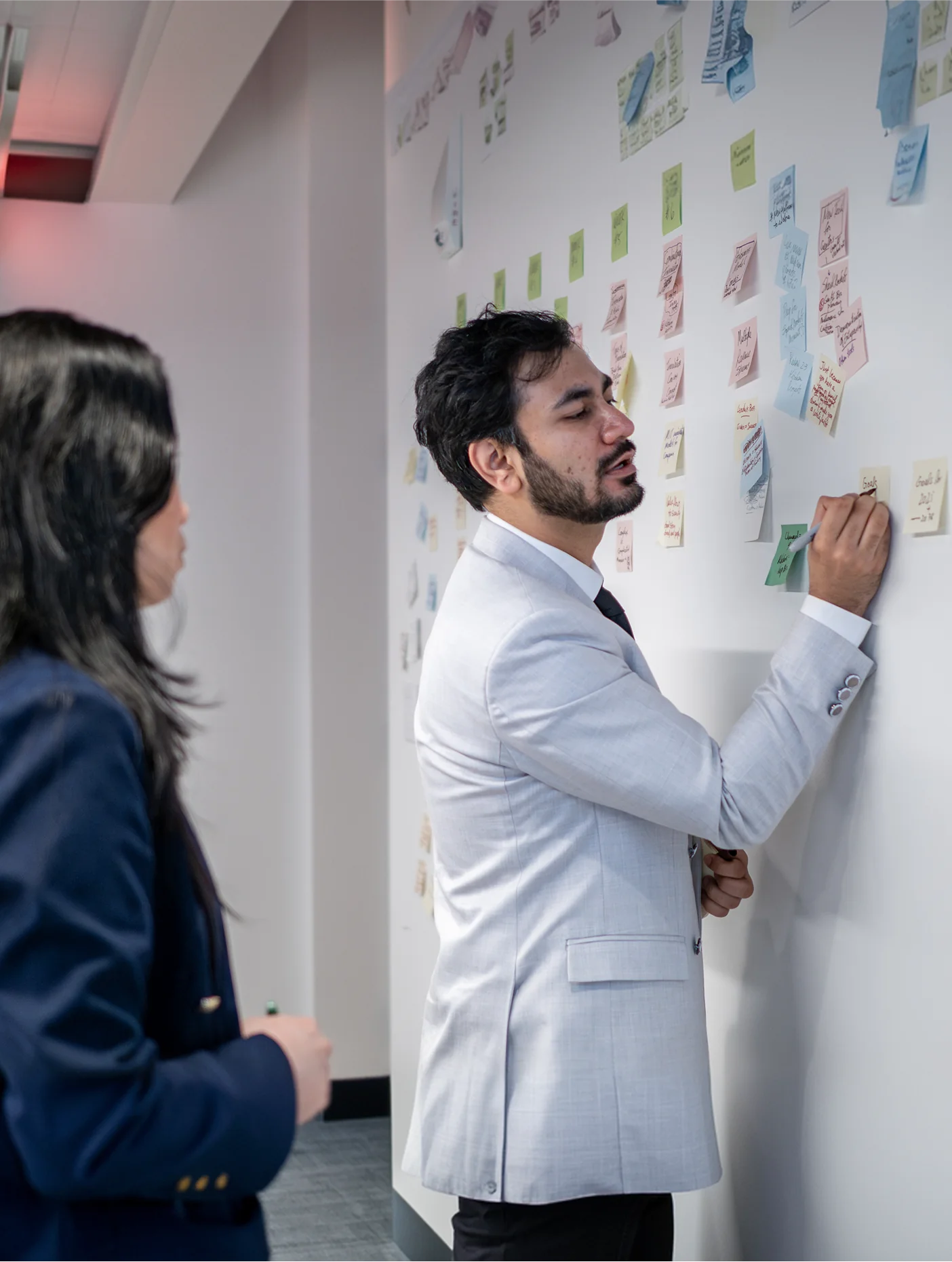 A man in a light suit writing on sticky notes on a wall while a woman watches
