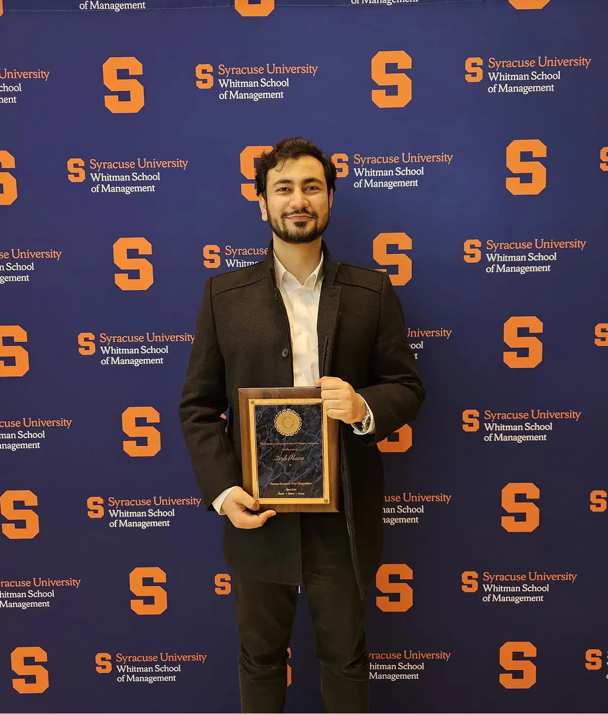 A man holding an award plaque in front of a Syracuse University Whitman School of Management backdrop