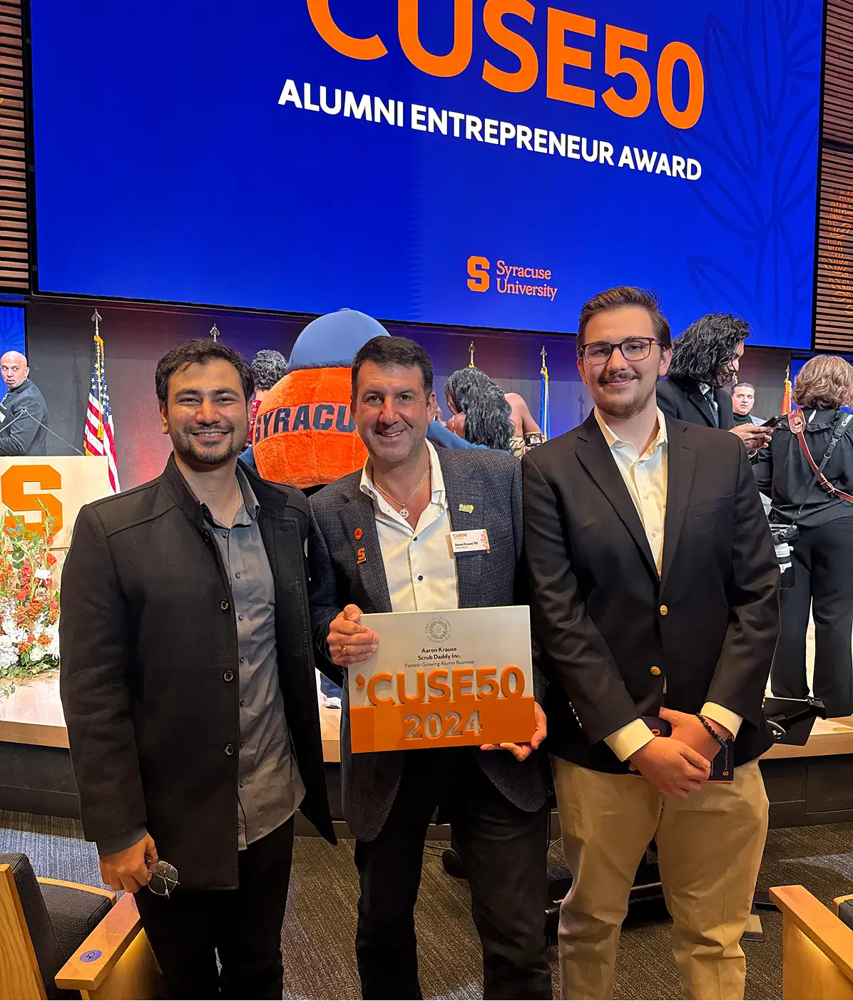 Three men smiling with a CUSE50 2024 award under a screen reading CUSE50 Alumni Entrepreneur Award, Syracuse University