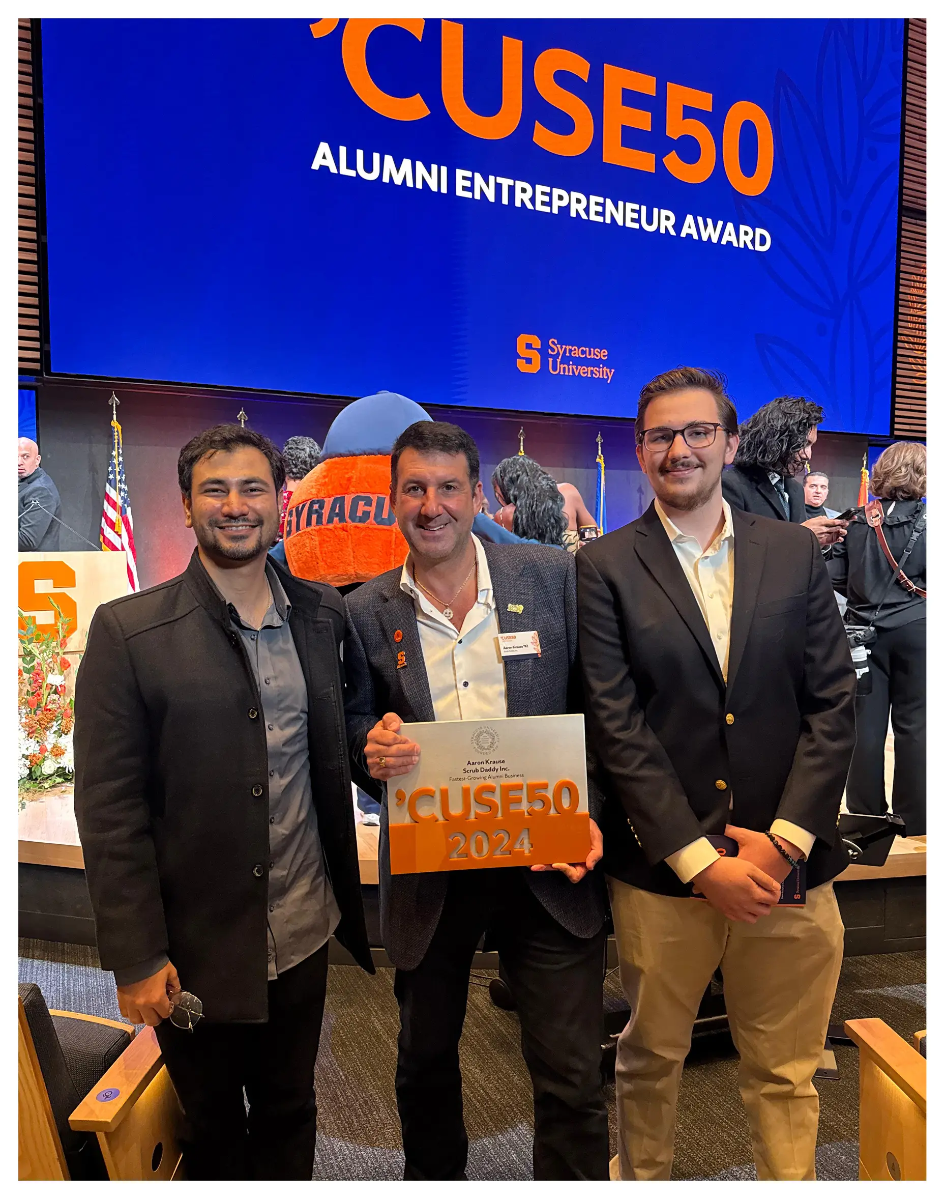 contact us Three men standing in front of a large screen with the words 'CUSE50 ALUMNI ENTREPRENEUR AWARD' on it, holding a