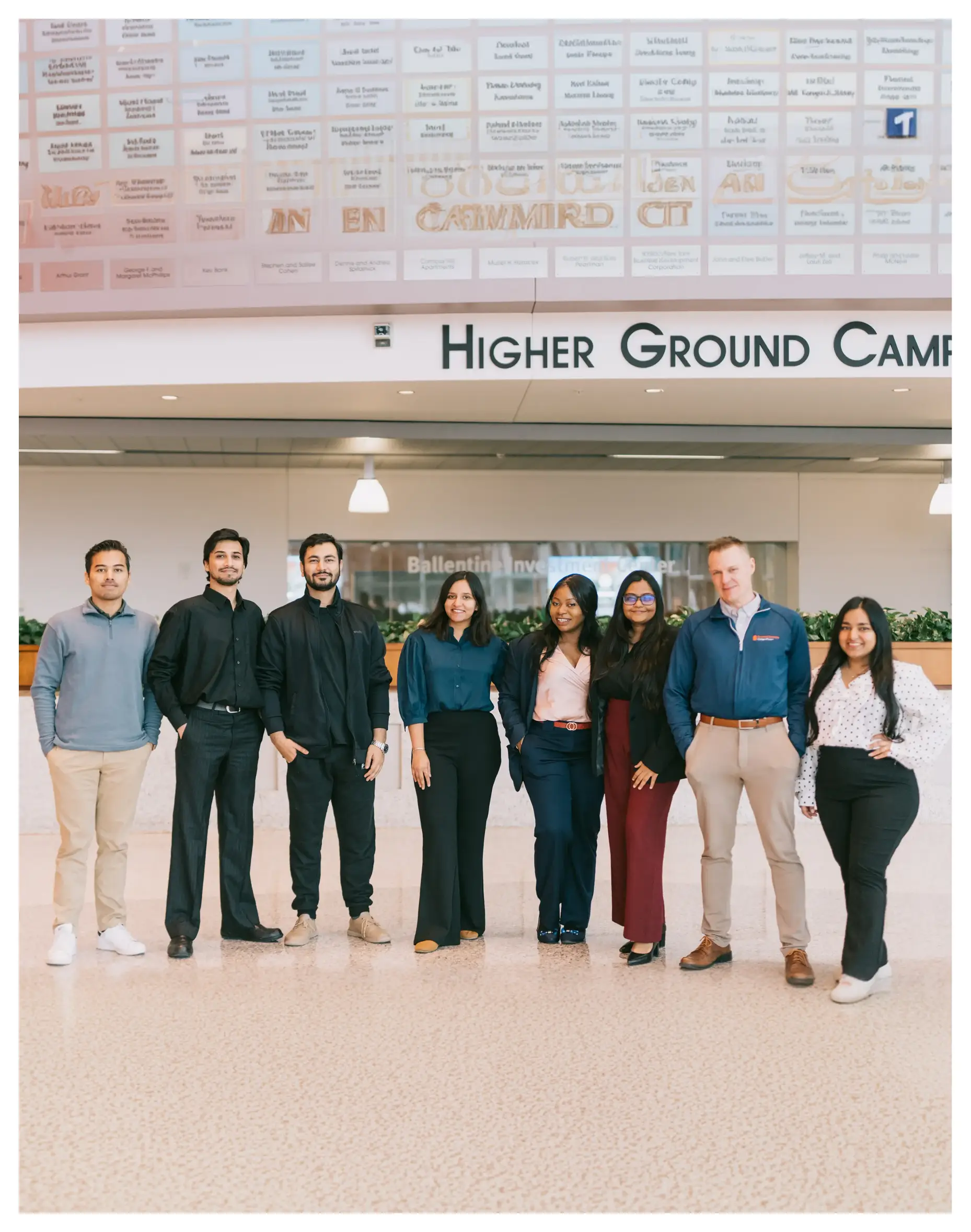 contact us A group of people standing in front of a sign that says 'Higher Ground Camp' and a wall with names on it