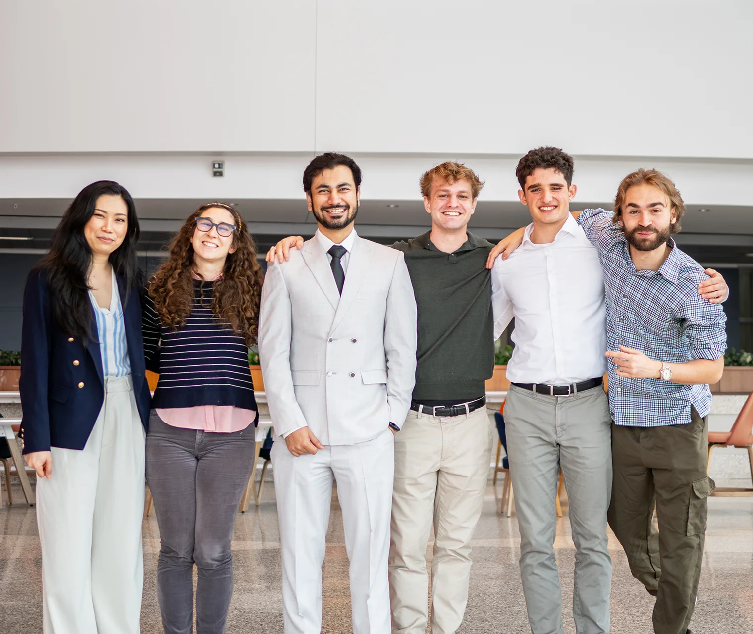 Six people standing arm in arm and smiling in a bright university hall