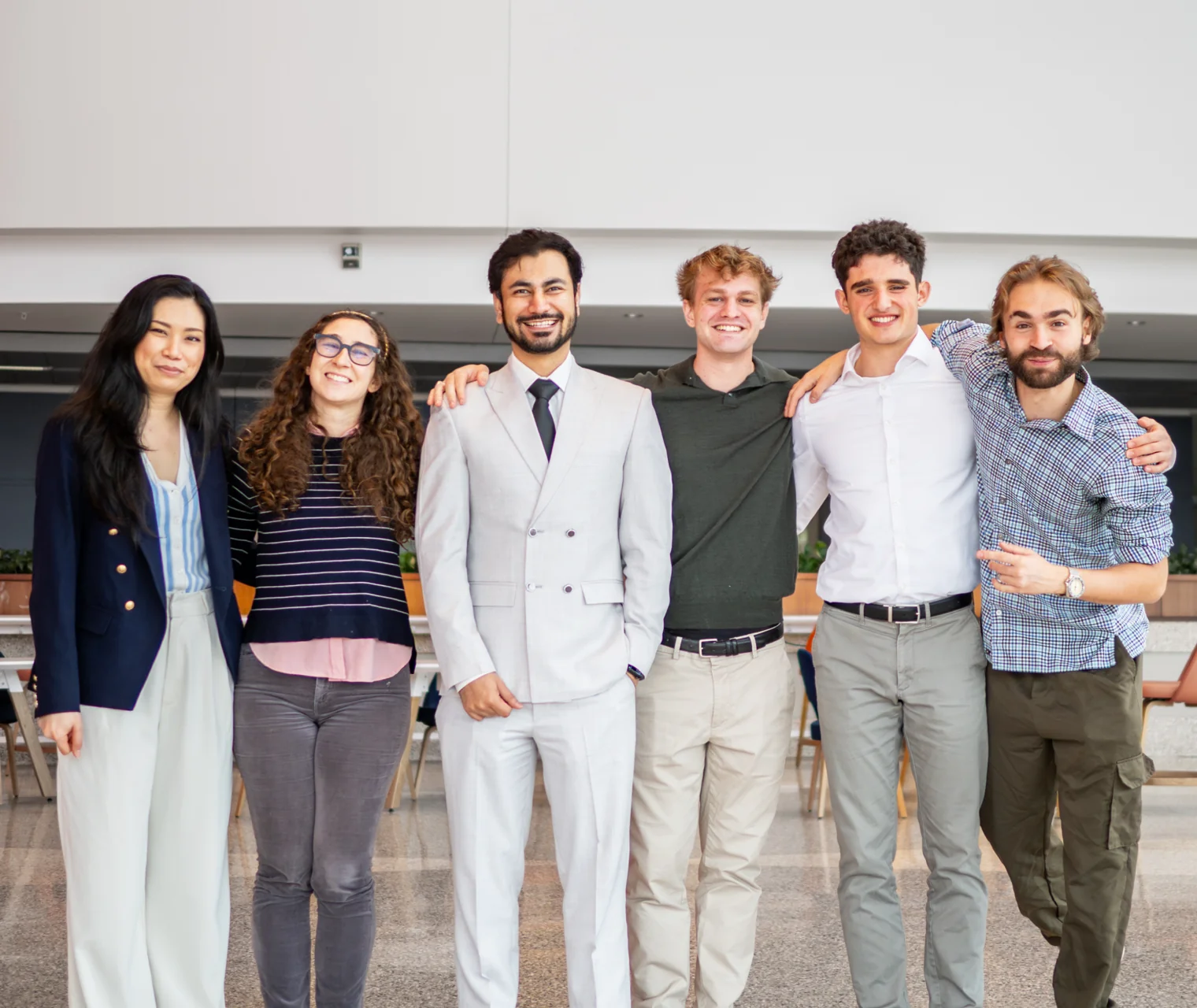 Six people standing arm in arm and smiling in a bright university hall