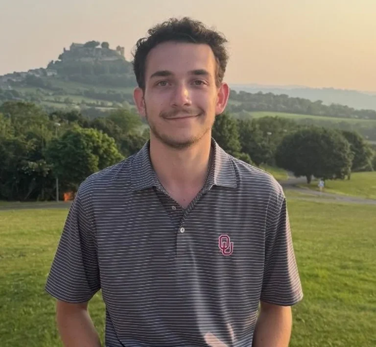 A young man in a striped polo shirt smiling outdoors on a grassy hill at sunset