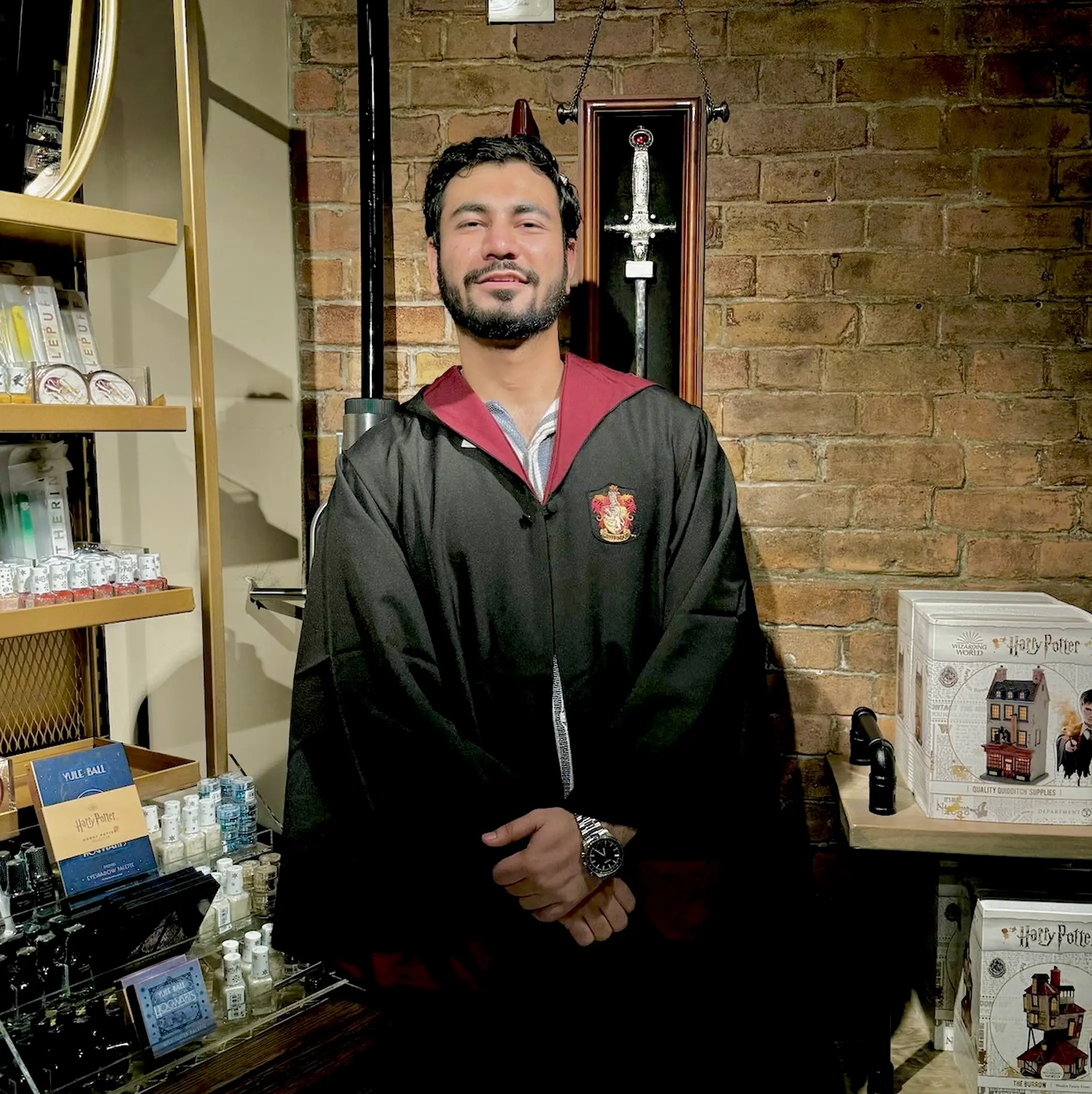 A smiling man in a Gryffindor style robe in a shop with a brick wall and Harry Potter boxes