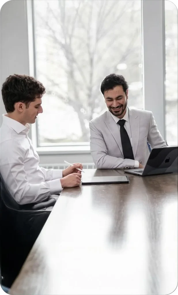 meeting Two men in business attire sitting at a table, smiling and looking at a laptop screen